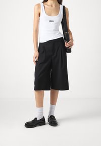 Kvinna i en vit ärmlös topp, svarta vida knälånga shorts, svarta loafers med vita strumpor och bär en svart väska över ena axeln.