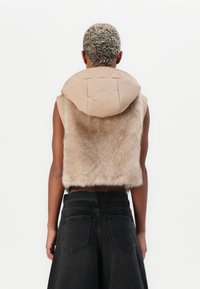 Gilet in finta pelliccia beige con cappuccio trapuntato beige, caratterizzato da un design senza maniche e un taglio corto. Jeans neri a vita alta con tasche posteriori.
