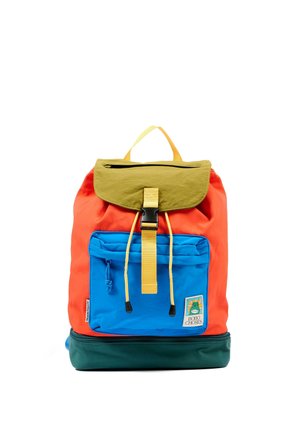 Zaino colorato con patta oliva, cinturino giallo, corpo arancione, tasca frontale blu e base verde scuro, con una piccola toppa sulla tasca.
