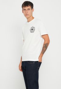 Un tricou alb din bumbac cu imprimeu logo pe piept, mâneci scurte și guler clasic tip crew, asortat cu blugi denim de un albastru închis.