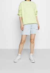 Sudadera oversized de color verde claro, pantalones cortos de mezclilla azul con dobladillo deshilachado y un desgarro, combinados con zapatillas blancas de caña alta.