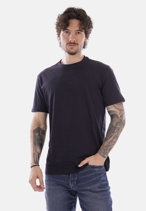 Uomo con capelli ricci e scuri e tatuaggi su entrambi gli avambracci, indossa una maglietta nera e jeans blu, in piedi con una mano in tasca.