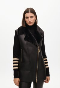 Chaleco de cuero negro con cuello de piel, con cremallera dorada, bolsillos laterales y acentos a rayas en las mangas. Textura suave.