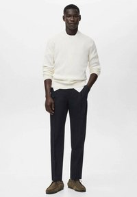 Homme debout les mains dans les poches, portant un pull blanc à côtes, un pantalon noir et des chaussures en daim marron à lacets sur fond blanc.