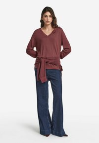 Pull de couleur bordeaux à col en V avec des manches longues, doté d'un détail de bandeau noué à la taille, associé à un jean bleu foncé à jambes larges. Texture lisse dans l'ensemble.