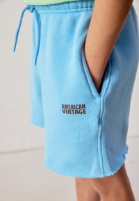 Personne portant un short à cordon de serrage bleu clair avec l'inscription « AMERICAN VINTAGE », main reposant dans la poche latérale, fond beige.