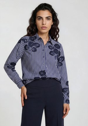 Frau mit dunklen, welligen Haaren, die ein marineblau-weiß gestreiftes Hemd mit Knöpfen und Blumenmuster sowie dunkelblaue Hosen trägt, steht vor einem einfarbigen Hintergrund.