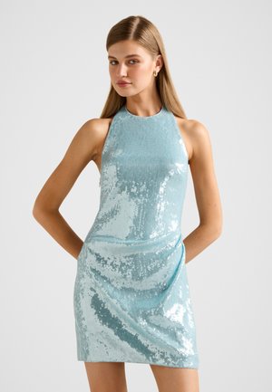 Kobieta w bezrękawowej, jasnoniebieskiej, cekinowej sukience mini na szyję, stojąca z rękami za plecami na neutralnym tle.