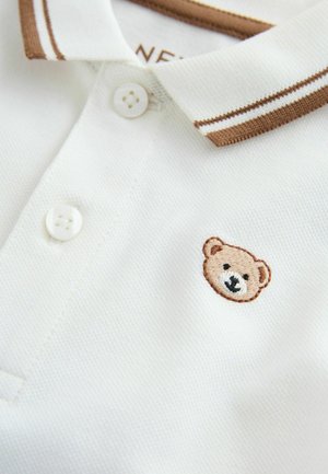 Wit poloshirt met een bruine geribbelde kraag en twee knopen. Heeft een geborduurd berengezicht aan de linkerkant. Zachte, gestructureerde stof.