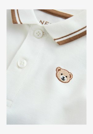 Bijela polo majica s smeđim rebrastim ovratnikom i dva gumba. Na lijevoj strani nalazi se izvezeni medvjeđi lik. Mekana, teksturirana tkanina.
