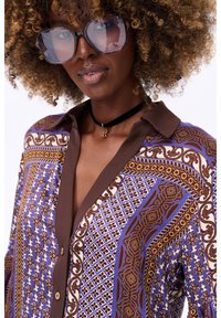 Multikleurige blouse met complexe patronen in blauw, bruin en wit. Kenmerkend is de bruine kraag en knopen. Gelaagde stof met een losse pasvorm.