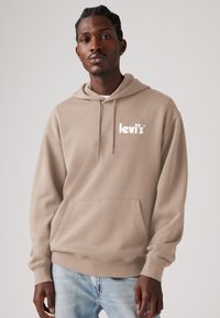 Beige hoodie met trekkoord, voorzak en wit logo. Zachte textuur, relaxte pasvorm. Draagt over lichtblauwe spijkerbroek.