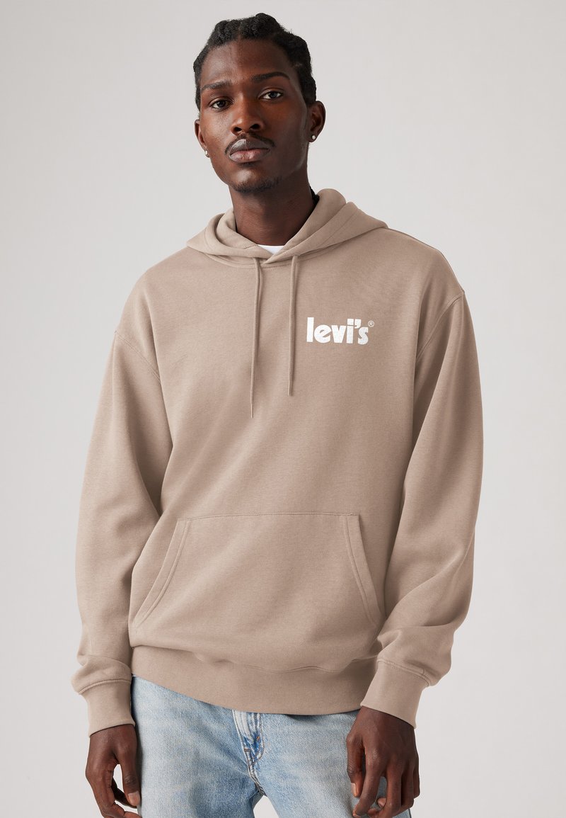 Haut à capuche beige avec cordon de serrage, poche avant et logo blanc. Texture douce, coupe décontractée. Porté par-dessus un jean bleu clair.