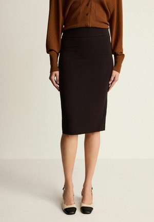 Jupe crayon noire en tissu extensible, dotée d'une taille haute et d'une finition lisse. Associée à un cardigan marron et à des talons bicolores.