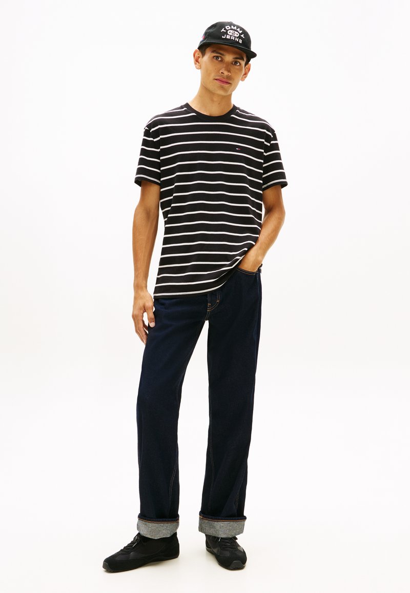 Jeune homme portant un t-shirt rayé noir et blanc, un jean bleu foncé retroussé, des baskets noires et une casquette noire, debout avec une main dans la poche.