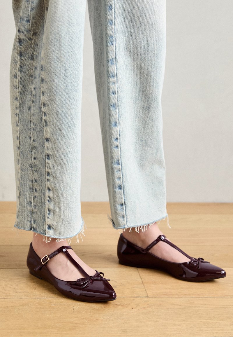 Pantofole ballerine in vernice borgogna con T-strap, punta affusolata e un piccolo dettaglio a fiocco, abbinate a jeans azzurri con orlo sfrangiato.