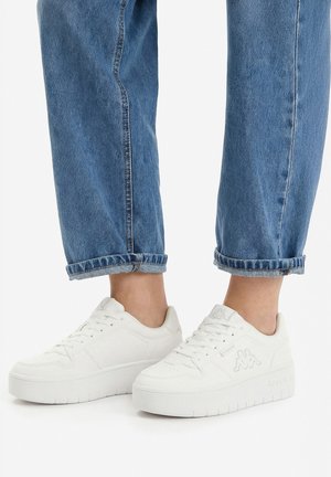 Osoba nosząca podwinięte niebieskie dżinsy i białe sneakersy na platformie z logo po bokach, stojąca na białej powierzchni.
