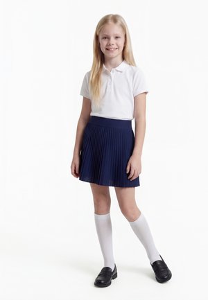 Uśmiechnięta dziewczyna z długimi blond włosami, ubrana w białą koszulkę polo, granatową plisowaną spódnicę, białe skarpetki do kolan i czarne buty, stojąca na białym tle.