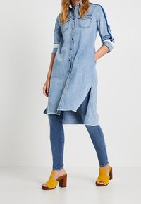 Ljusblå denimklänning med lång ärm, framknäppning, sidospår, i kombination med blå skinnyjeans och gula platåskor.