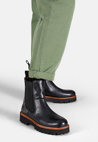 Sorte læder chelsea støvler med elastiske sidepaneler, robuste såler og orange detaljer langs sålen, parret med rullede olivengrønne bukser.