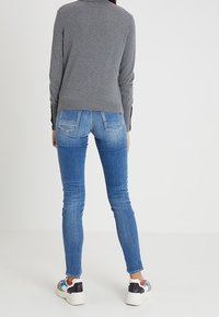 Person in einem grauen Pullover, ausgebleichten blauen Skinny-Jeans und mehrfarbigen Turnschuhen, steht mit dem Rücken zu einer einfarbigen Hintergrund.