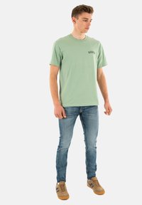 Lichtgroene t-shirt met korte mouwen met het "Dickies" logo, gecombineerd met vervaagde blauwe jeans en bruine sportschoenen met donkere strepen.