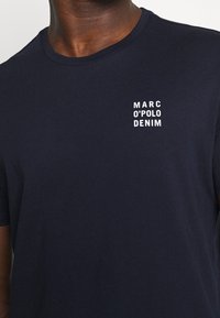 Tummansininen puuvillapaita, jossa on pyöreä kaula-aukko ja vasemmassa rinnassa valkoinen logo-teksti "MARC O'POLO DENIM". Kiiltävä pinta.