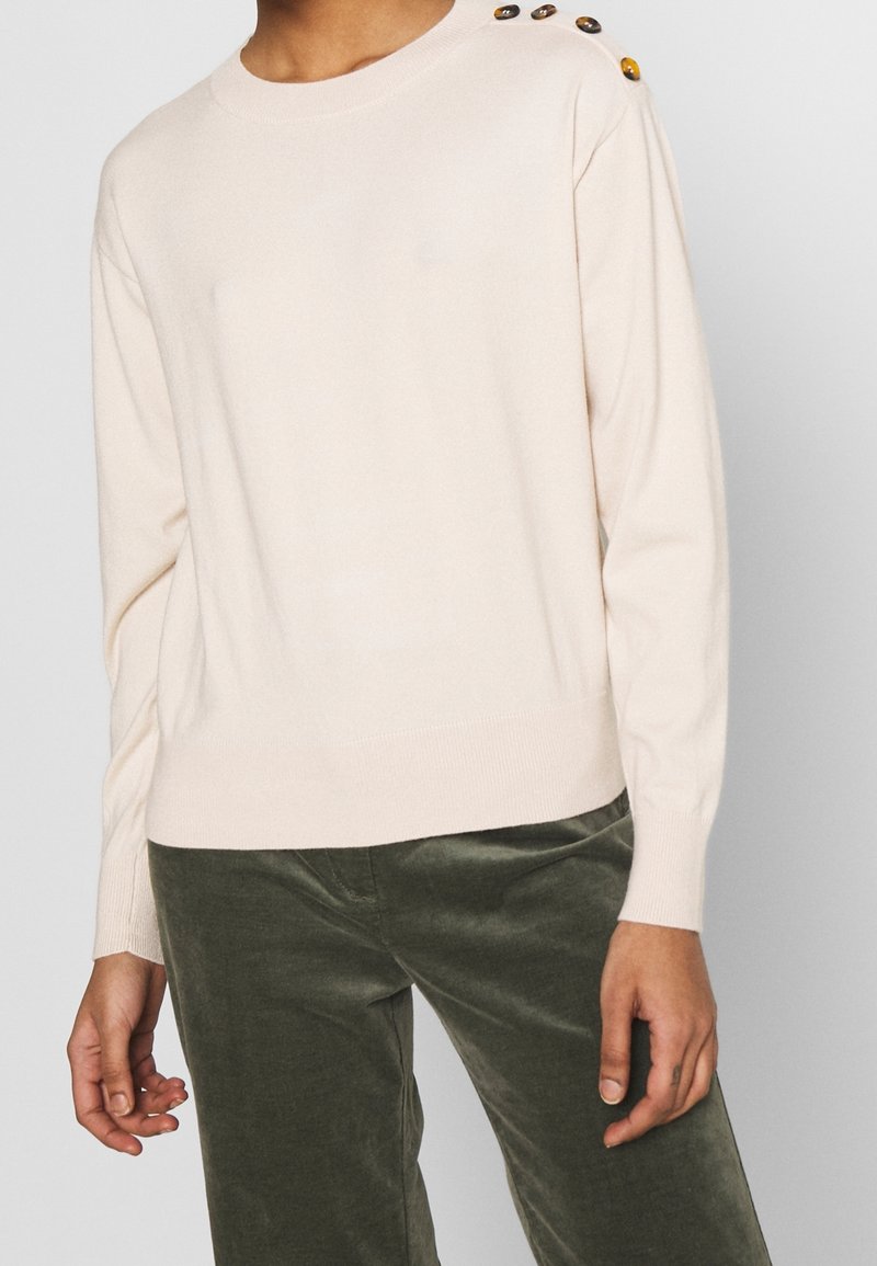 Pull crème à manches longues avec trois boutons décoratifs sur l'épaule gauche, porté avec un pantalon en velours vert foncé.