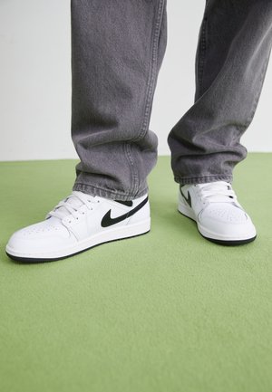 Biele kožené tenisky s čiernymi akcentmi, perforovaný špička, ploché šnúrky, okrúhla gumová podrážka a viditeľné logo Nike na stranách.