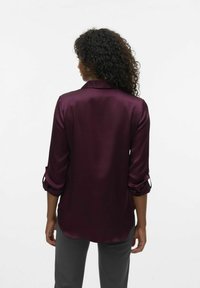 Chemise en satin bordeaux avec un col classique, des manches longues retroussées aux poignets et un ourlet arrondi. Associée à un pantalon gris.