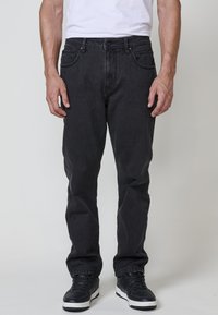 Hombre vestido con jeans rectos negros, camiseta blanca y zapatillas negras con suelas blancas, de pie contra un fondo gris liso.