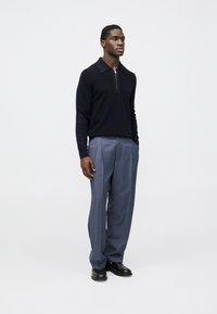 Námornícky modrý pulóver so zapínaním na zips a golierom, skombinovaný so svetlosivými nohavicami na mieru. Oblečenie má čisté línie a hladkú textúru. Čierne topánky.