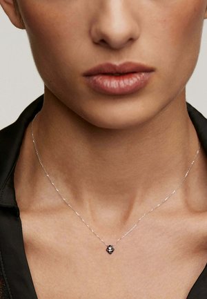 Collar de cadena de plata con un pequeño colgante en forma de diamante que presenta un acento oscuro. El fondo es neutro, centrándose en el collar.