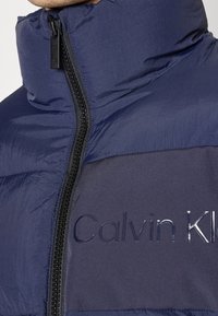 Mörkblå puffjacka med hög krage, med en svart dragkedja och präglad "Calvin Klein" logotyp på bröstet i ett kontrasterande mörkt tyg.