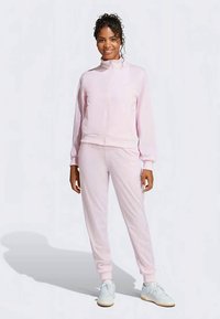 Ženska stoji, oblečena v svetlo roza zip-upp track jakno in usklajene hlače jogger ter bele superge, ter se smeška v kameru.