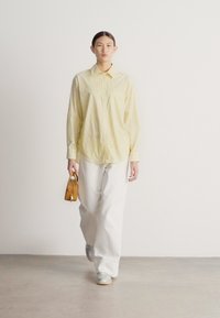 Dzeltens svītrains krekls ar pogām, brīvi krītošas baltas bikses, sudraba krāsas sporta apavi un maza brūna rokassoma. Modelis stāv pret vienkāršu fona sienu.