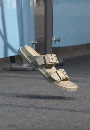 Sandalo in gomma beige con due fibbie regolabili che fluttua sopra una superficie asfaltata con pannelli blu sullo sfondo.