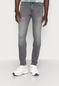 Jean gris slim avec une finition légèrement délavée, design à cinq poches et détails subtilement cousus, associé à des chaussures de sport blanches.