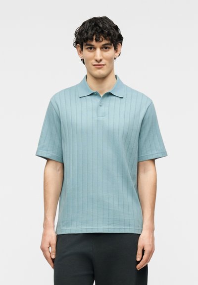 Jeune homme aux cheveux courts et bouclés portant un polo bleu clair à manches courtes côtelé et un pantalon noir, debout devant un fond uni.