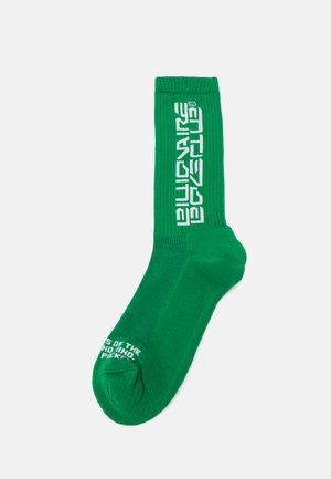 Chaussette en coton vert avec texture côtelée. Présente un texte logo blanc sur le côté et un texte blanc supplémentaire près de l'orteil. Longueur mi-mollet.