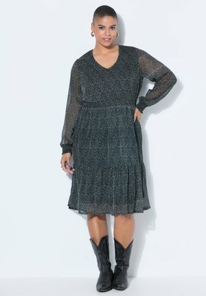 Kvinde iført en sort, mønstret knælængde kjole med gennemsigtige lange ærmer og sorte cowboystøvler, stående mod en hvid baggrund.