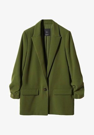 Persona che indossa un blazer verde oliva sopra una maglietta bianca, jeans blu a vita alta e dritti e scarpe nere appuntite, in piedi su un pavimento grigio.