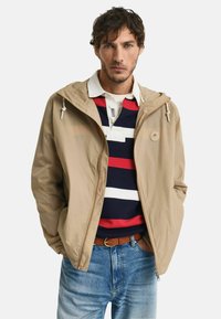 Giacca leggera beige con cappuccio, chiusura con zip e lacci, indossata sopra una polo a righe blu navy con accenti rossi e bianchi.