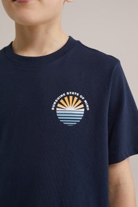 Granatowa bawełniana koszulka z okrągłym nadrukiem, przedstawiającym żółto-niebieskie słońce wschodzące nad niebieskimi i białymi paskami.