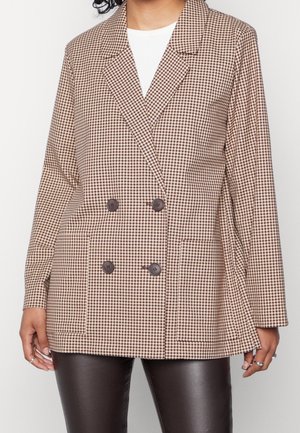 Frau trägt einen beige-braun karierten zweireihigen Blazer mit vier dunklen Knöpfen, über einem weißen Top und schwarzen Lederhosen.