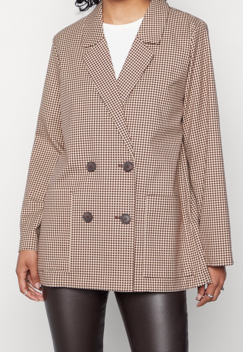 Femme portant un blazer croisé à carreaux beige et marron avec quatre boutons foncés, par-dessus un haut blanc et un pantalon en cuir noir.