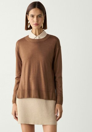 Kvinde med glat brunt hår bærer guldsmykker, en brun trøje over en beige skjorte med krave og en beige nederdel mod en ensfarvet baggrund.