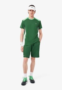 Îmbrăcăminte sport verde cu un top cu mânecă scurtă, având accente verde deschis și pantaloni scurți asortați. Include o visieră albă și pantofi sport verzi.