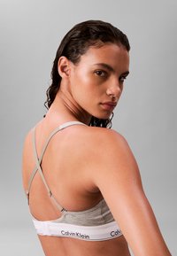 Jonge vrouw met nat haar die een grijze Calvin Klein beha met gekruiste bandjes draagt, kijkt over haar schouder tegen een effen grijze achtergrond.