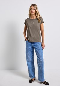 T-shirt vert olive à manches courtes avec un col rond, associé à un jean taille haute bleu clair à jambes larges. Des sandales tissées noires complètent la tenue.